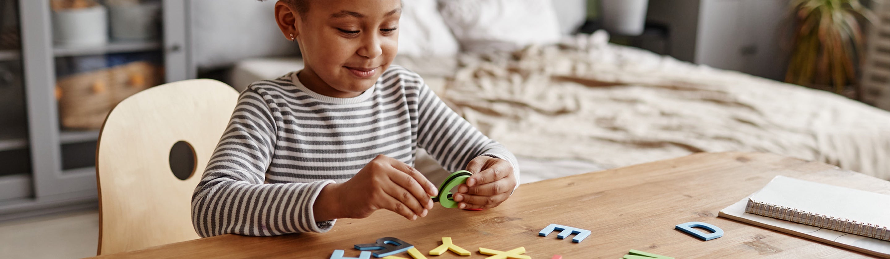 A Black child, about 4 years old, wearing a cozy striped shirt, looks at and studies large colorful letters and a desk.