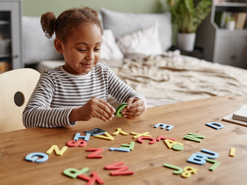 A young Black girl plays with colorful letters in a cozy home setting.